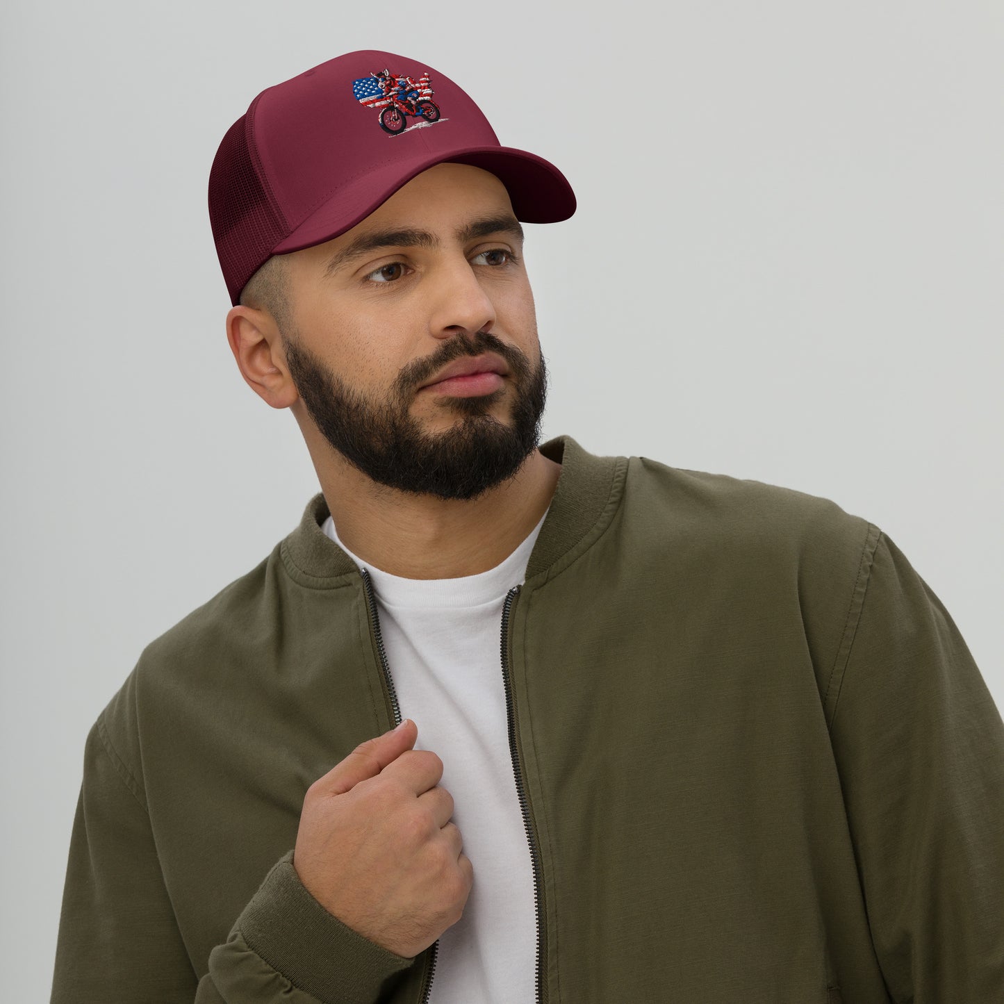 Man wearing a maroon cap with a design, olive green jacket, and white shirt on a plain background