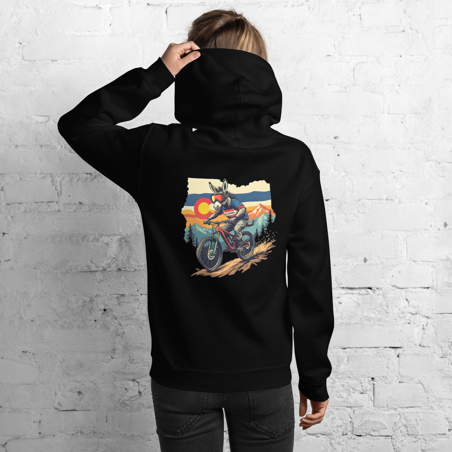 Person wearing a black hoodie with a graphic of a person on a motorcycle in a mountainous landscape on the back, against a white brick wall.