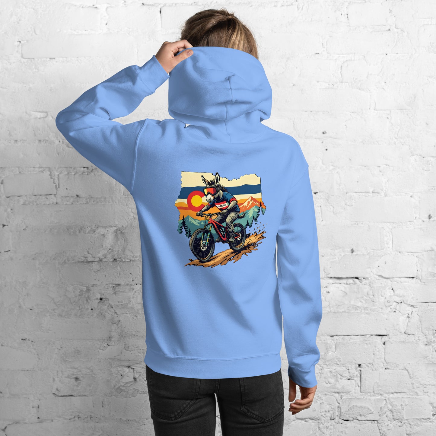Person wearing a light blue hoodie with a graphic of a motorcycle and mountains on the back, against a white brick wall.