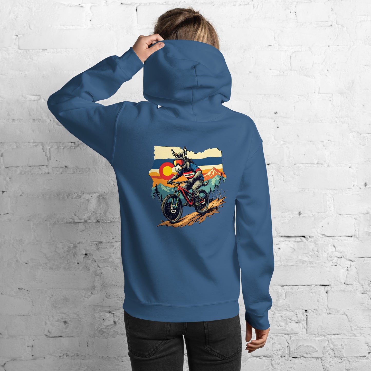 Person wearing a blue hoodie with a graphic of a person on a motorcycle and sunset landscape on the back, against a white brick wall.