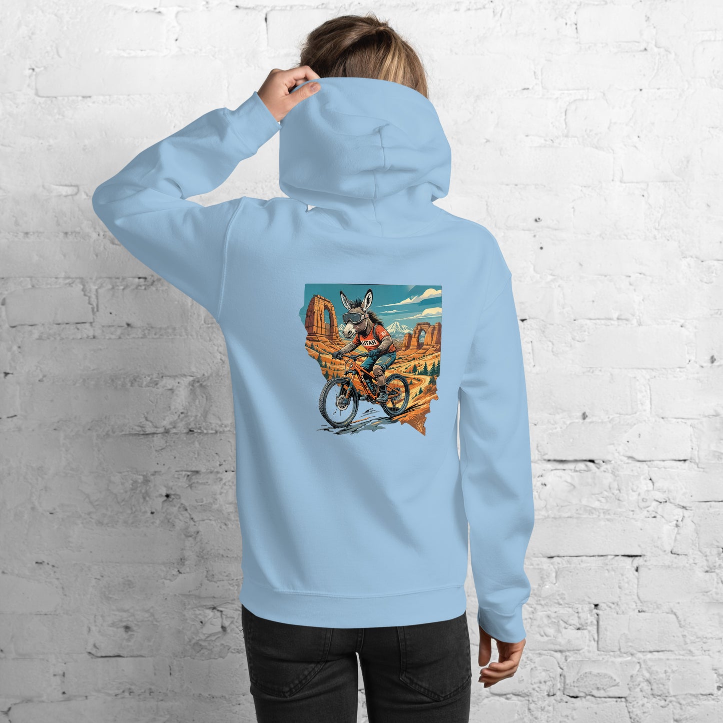 Person wearing a light blue hoodie with a graphic design on the back, standing against a white brick wall.