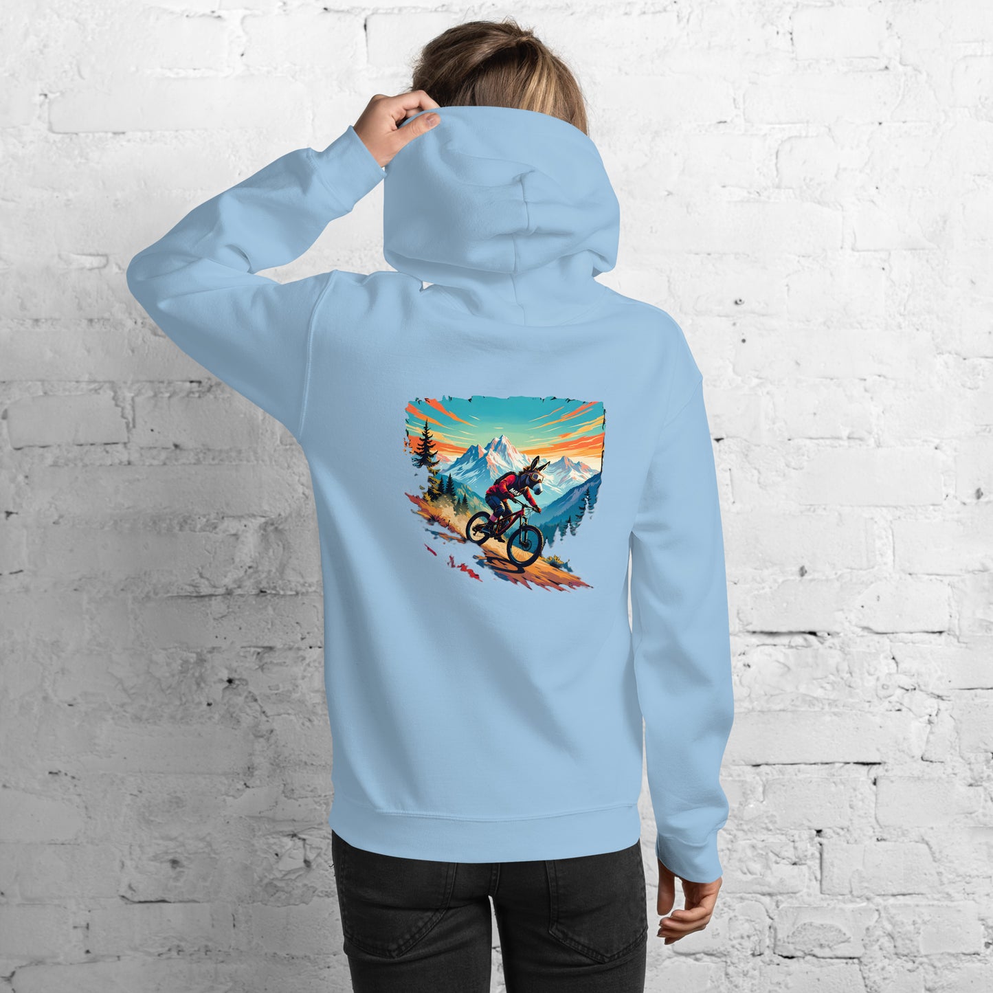 Light blue hoodie with a graphic of a person on a mountain bike in a scenic setting, worn by a person against a white brick wall.