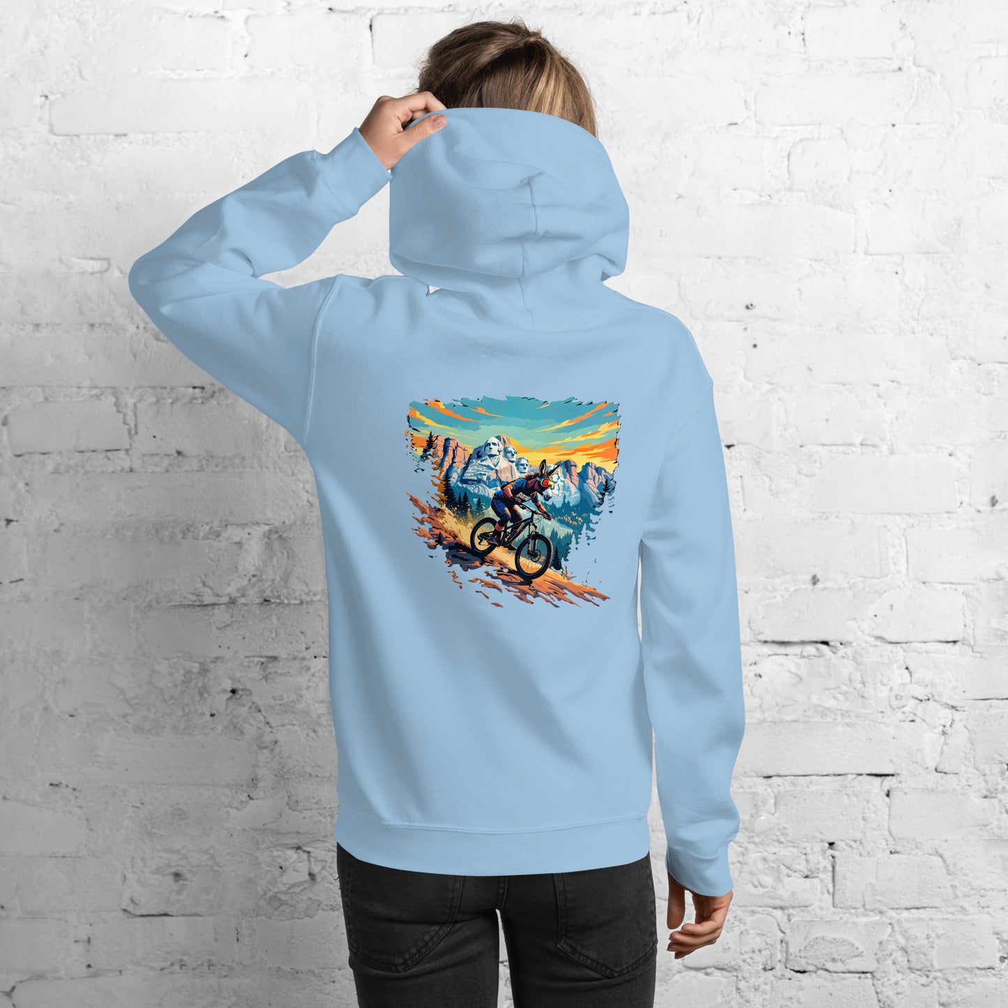Person wearing a light blue hoodie with a mountain bike graphic on the back against a white brick wall.