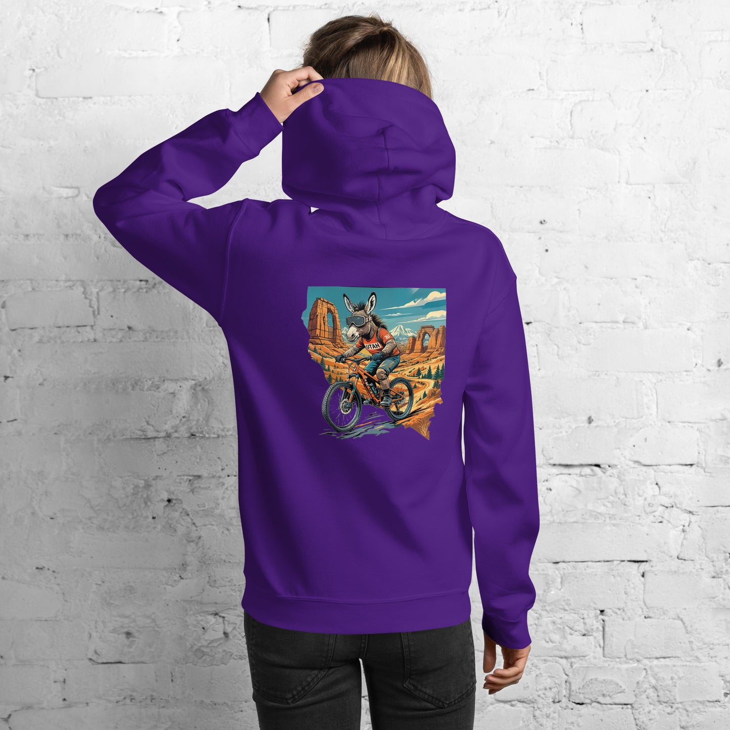 Person wearing a purple hoodie with a graphic design on the back against a white brick wall.