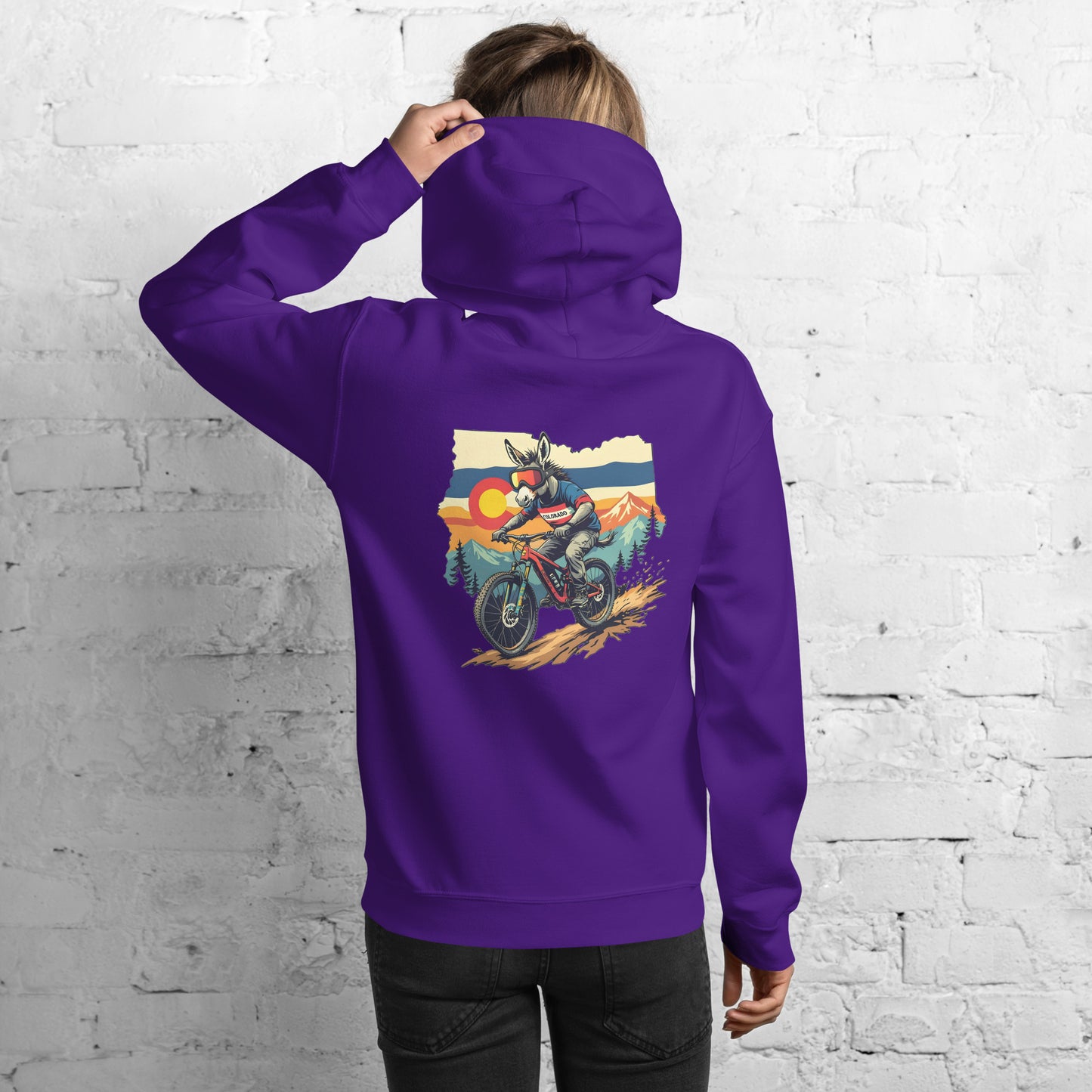 Person wearing a purple hoodie with a graphic design on the back, standing against a white brick wall.