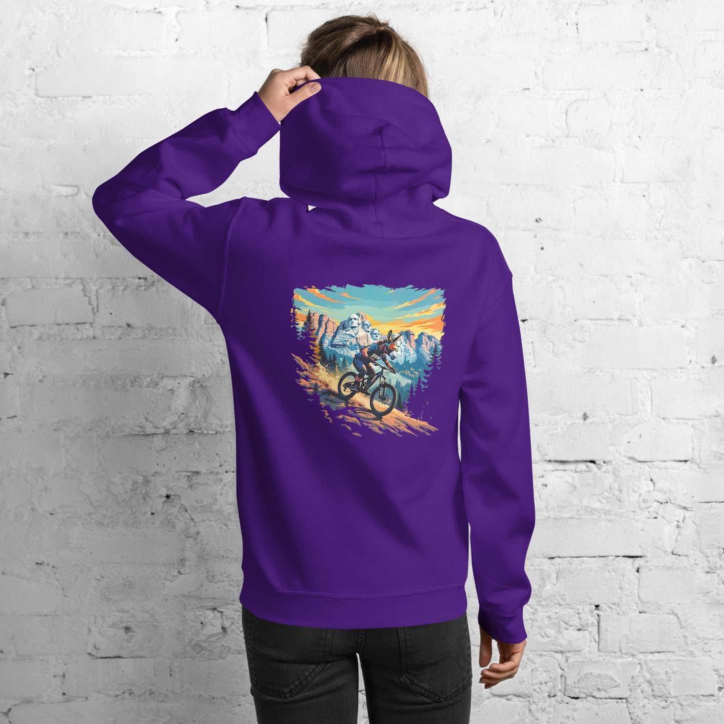 Person wearing a purple hoodie with a mountain bike graphic on the back, standing against a white brick wall.