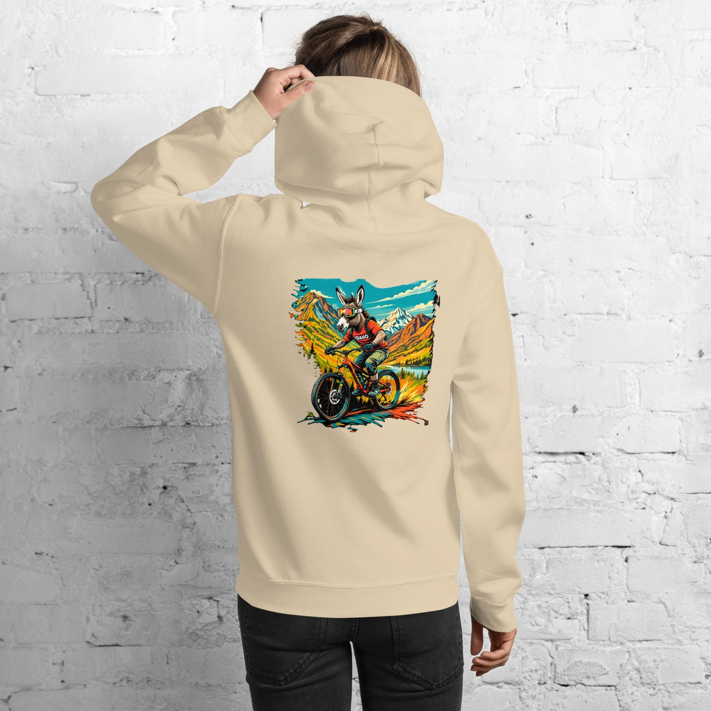 Person wearing a beige hoodie with a colorful graphic on the back against a white brick wall.