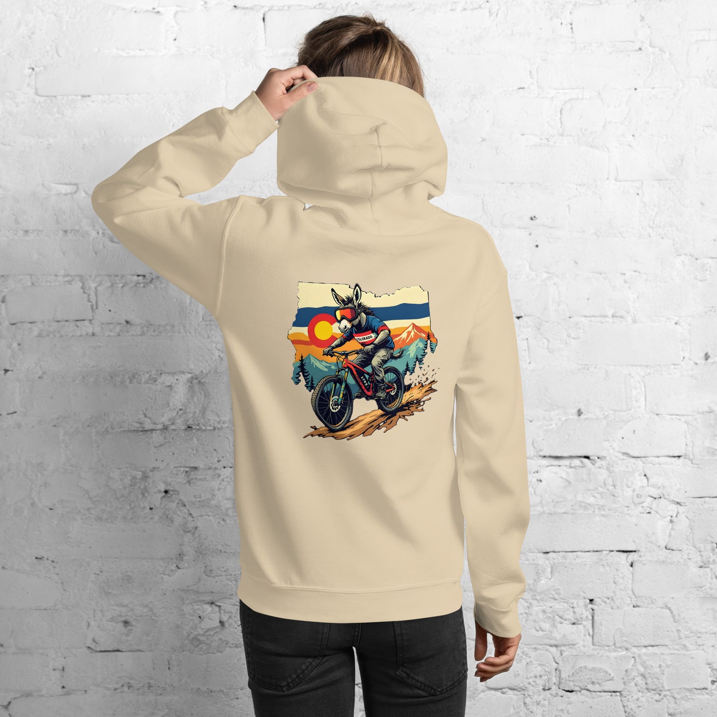 Person wearing a beige hoodie with a graphic of a motorcycle on the back against a white brick wall.