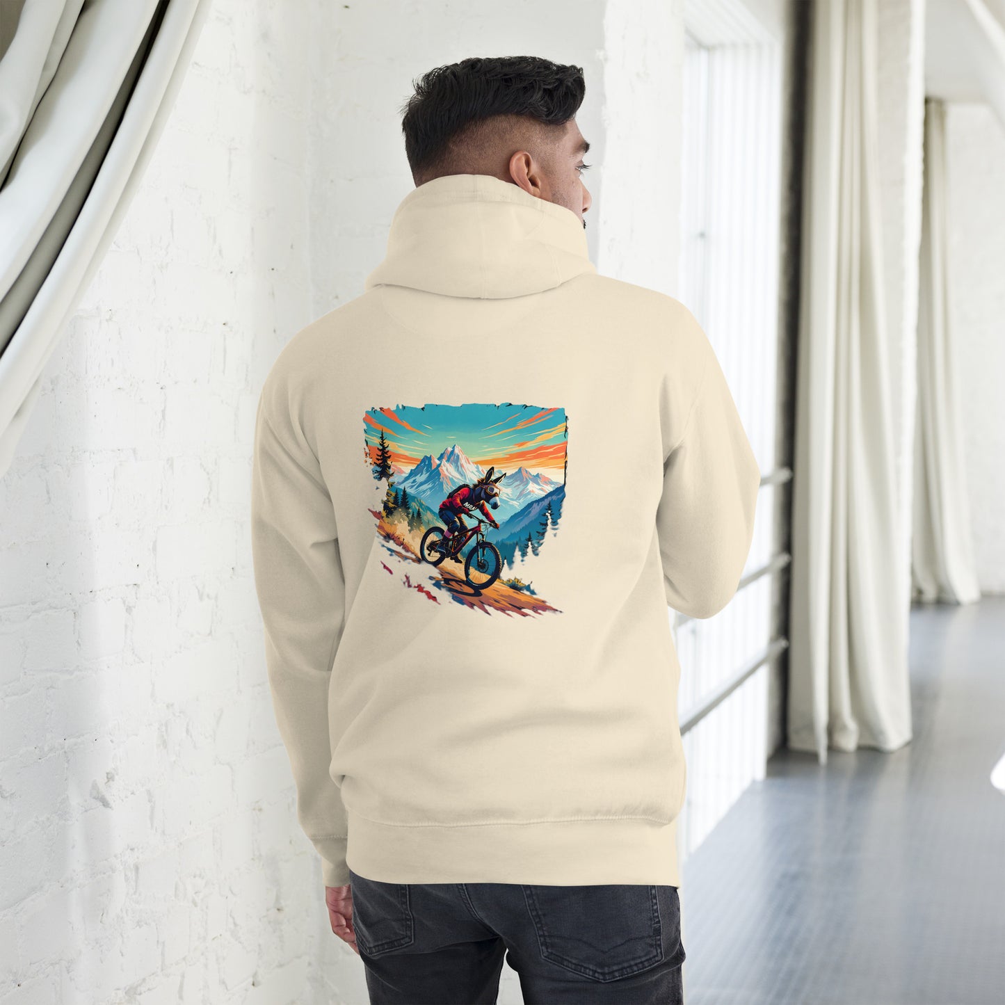 Person wearing a beige hoodie with a mountain bike graphic on the back, standing in a room with white walls and a window.