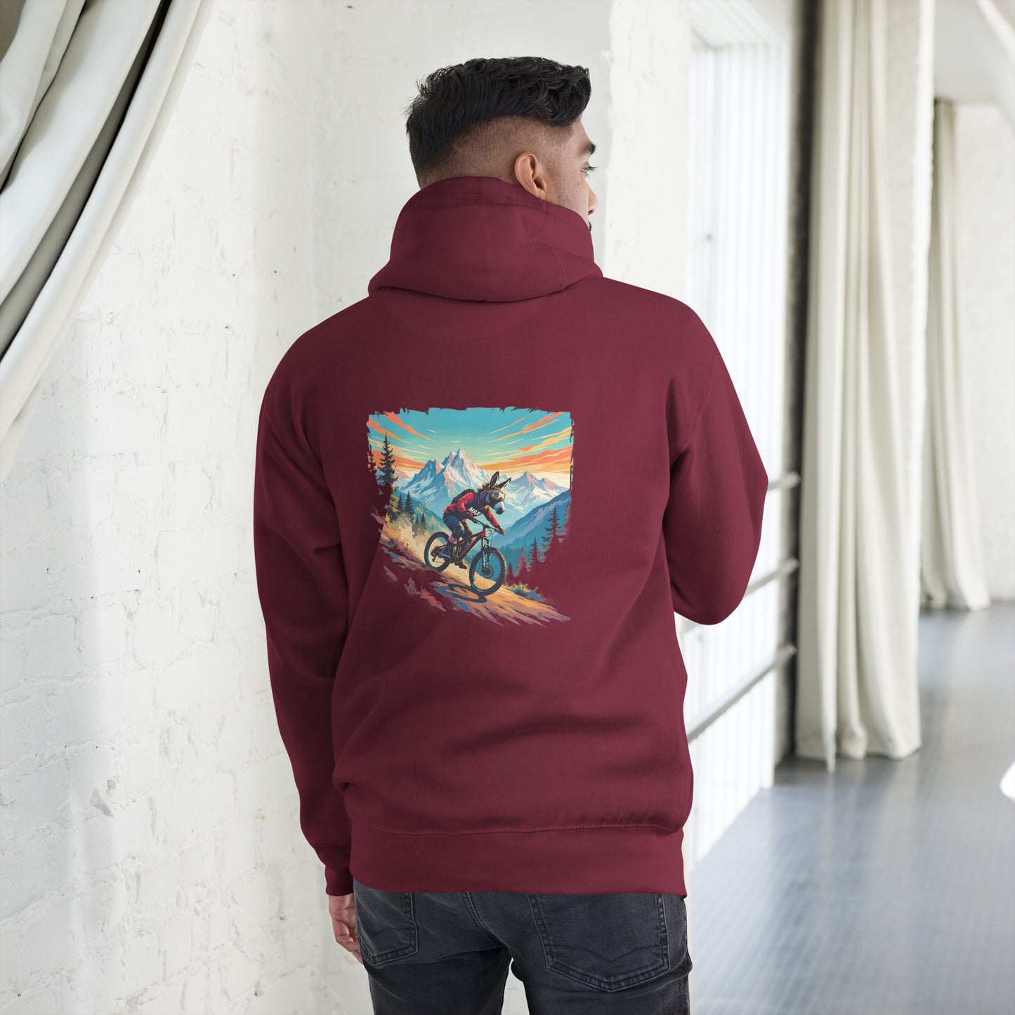 Person wearing a maroon hoodie with a graphic of a mountain and cyclist on the back, standing in a room with white walls and curtains.