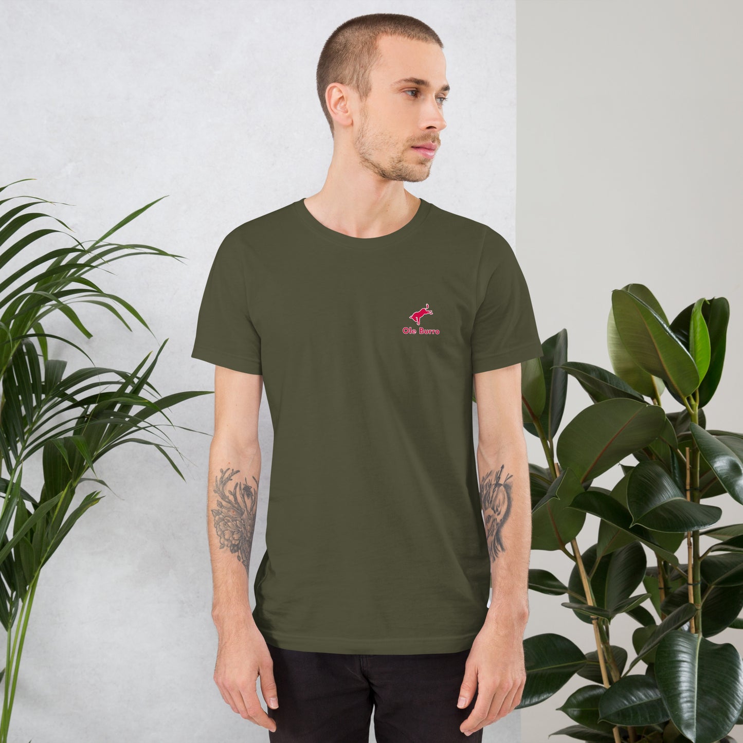 Man wearing a green t-shirt with a logo, standing between two plants against a light background
