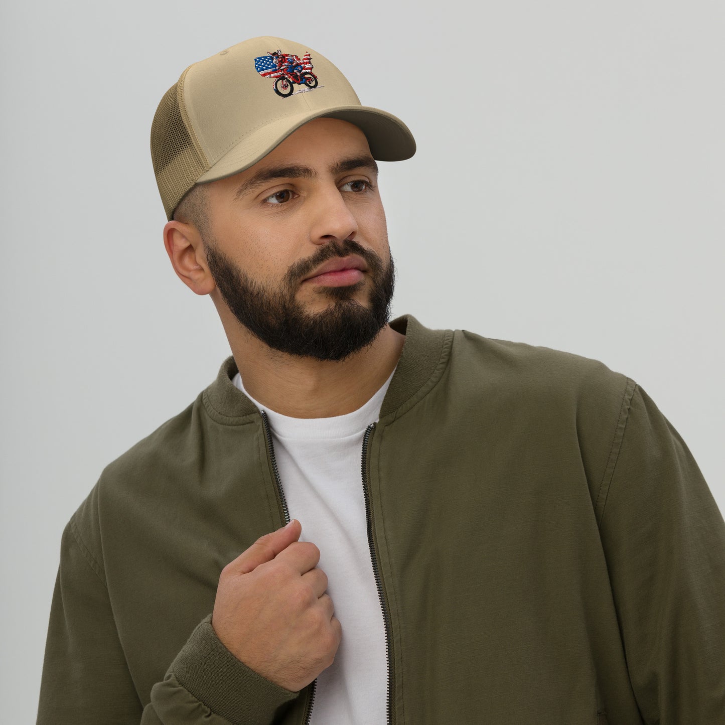 Man wearing a beige cap with an embroidered design and a green jacket over a white shirt.