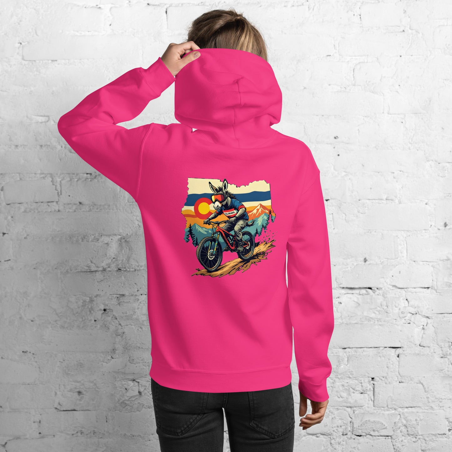 Person wearing a pink hoodie with a graphic of a person on a motorcycle in the mountains on the back, against a white brick wall.