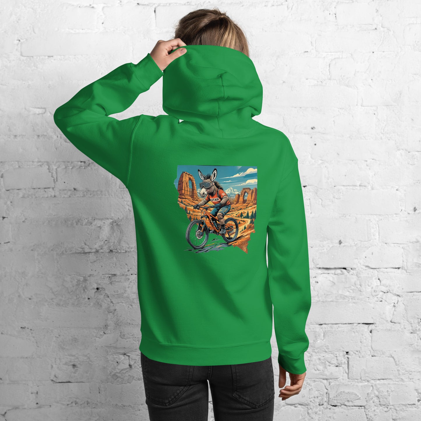 Person wearing a green hoodie with a graphic design on the back against a white brick wall.