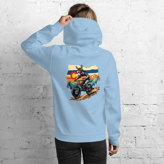 Person wearing a light blue hoodie with a graphic of a motorcycle on the back, standing against a white brick wall.