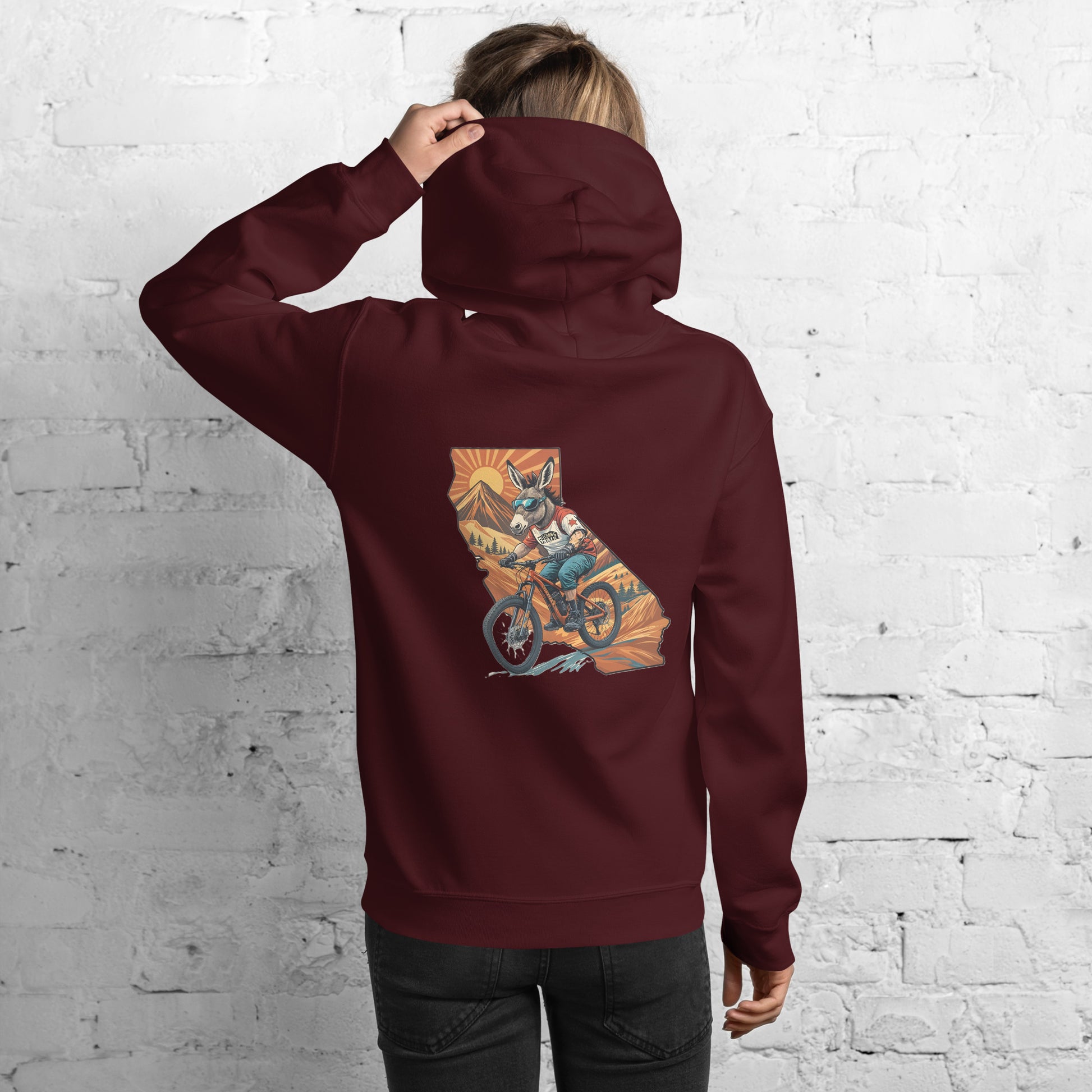 Person wearing a maroon hoodie with a graphic design on the back against a white brick wall.