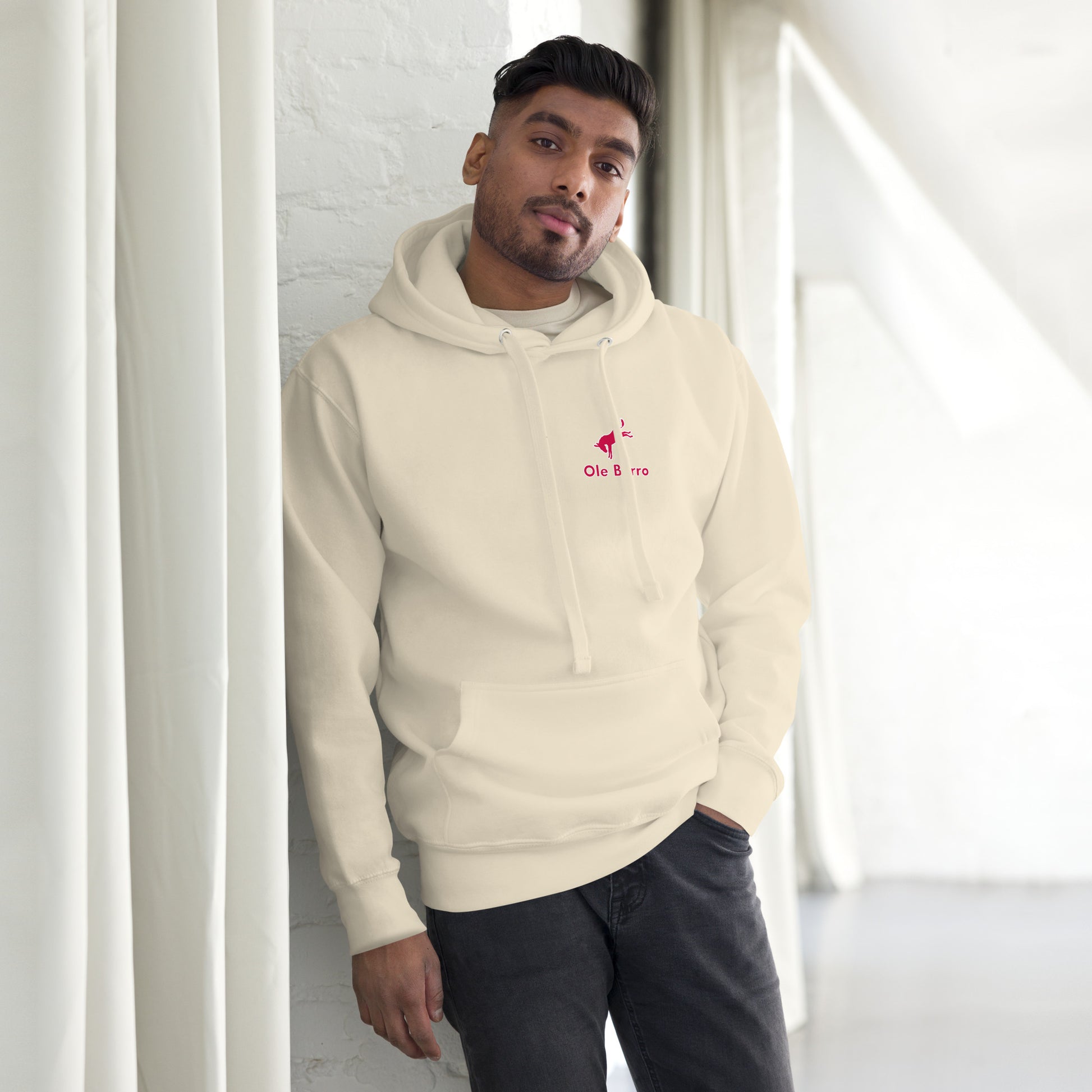 Man wearing a beige hoodie with a logo, standing against a white wall.
