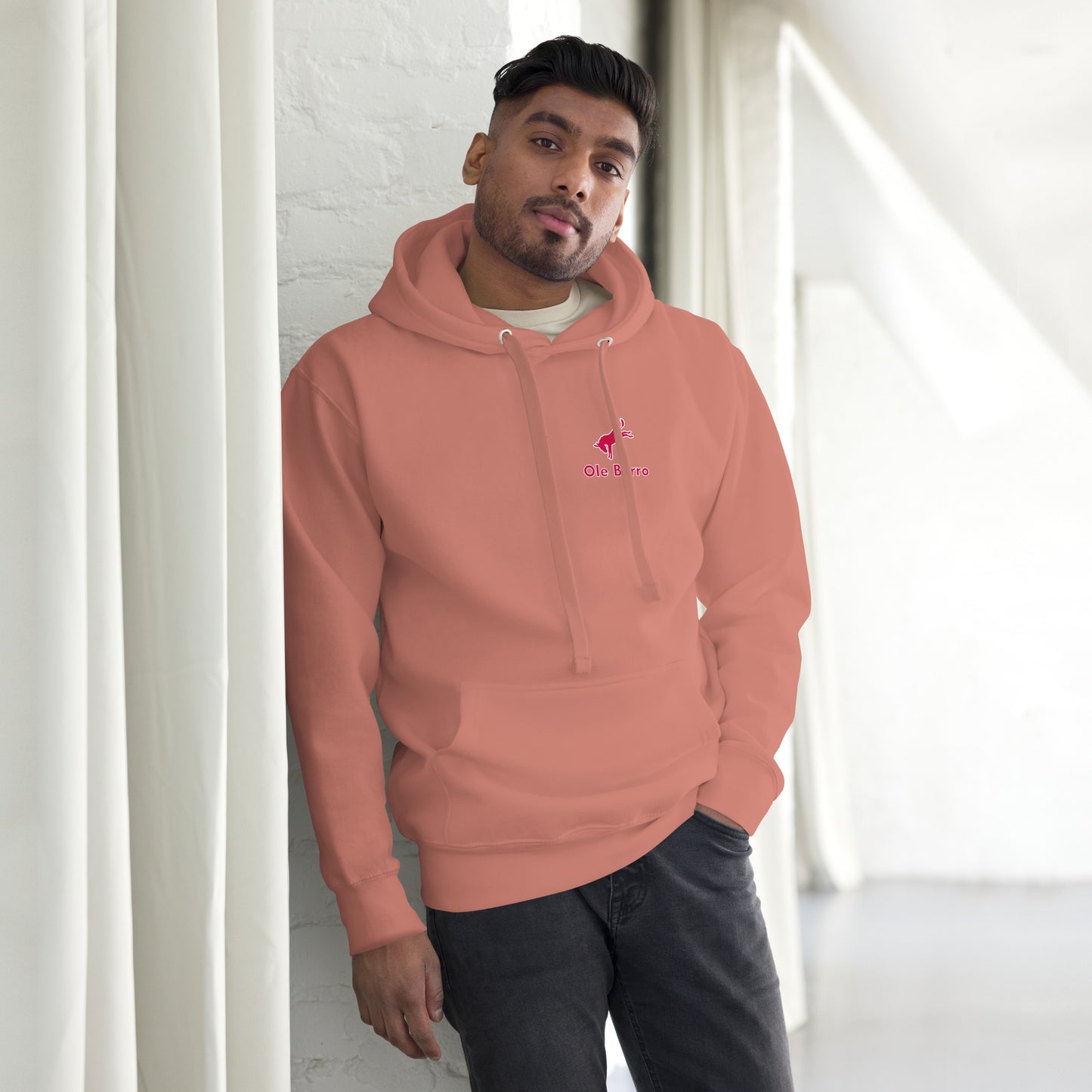 Man wearing a peach-colored hoodie with a logo, standing against a white wall.
