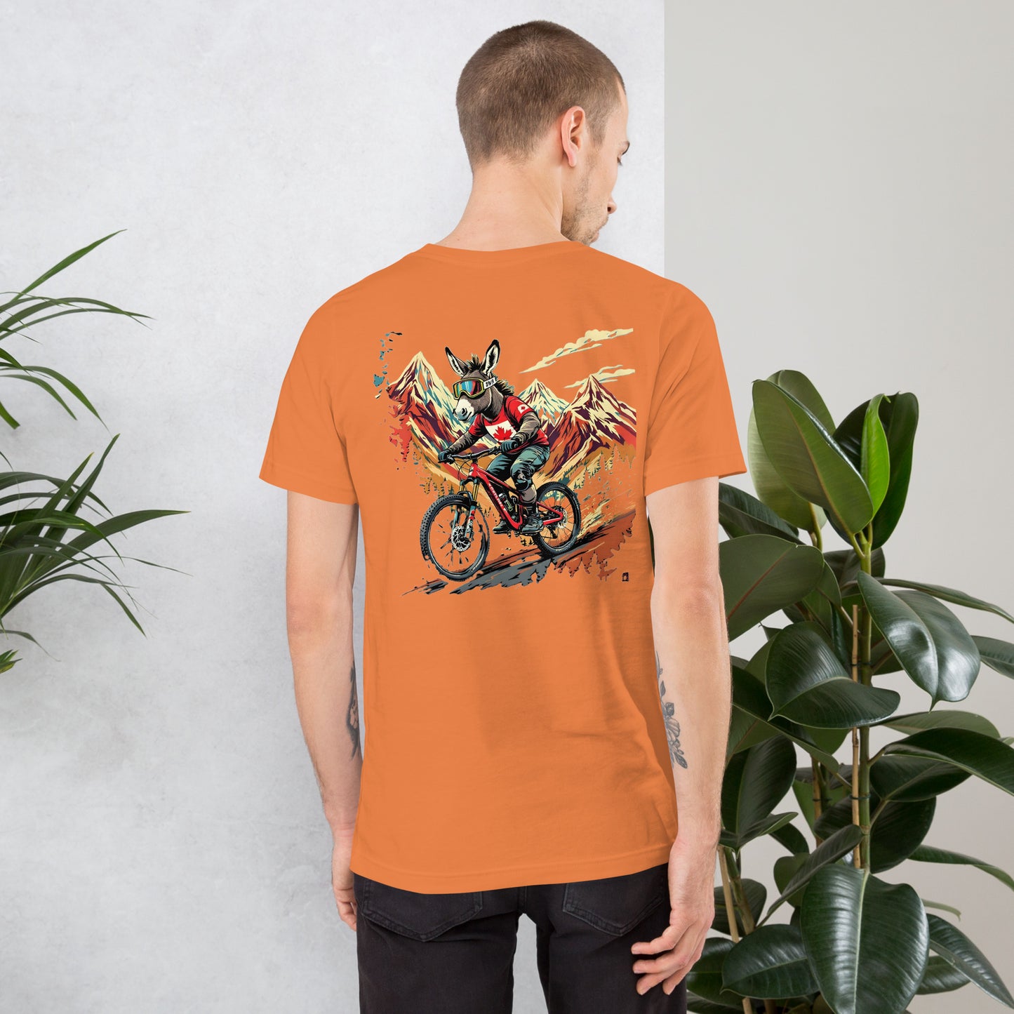 Person wearing an orange t-shirt with a graphic design on the back, standing against a light gray wall with plants.