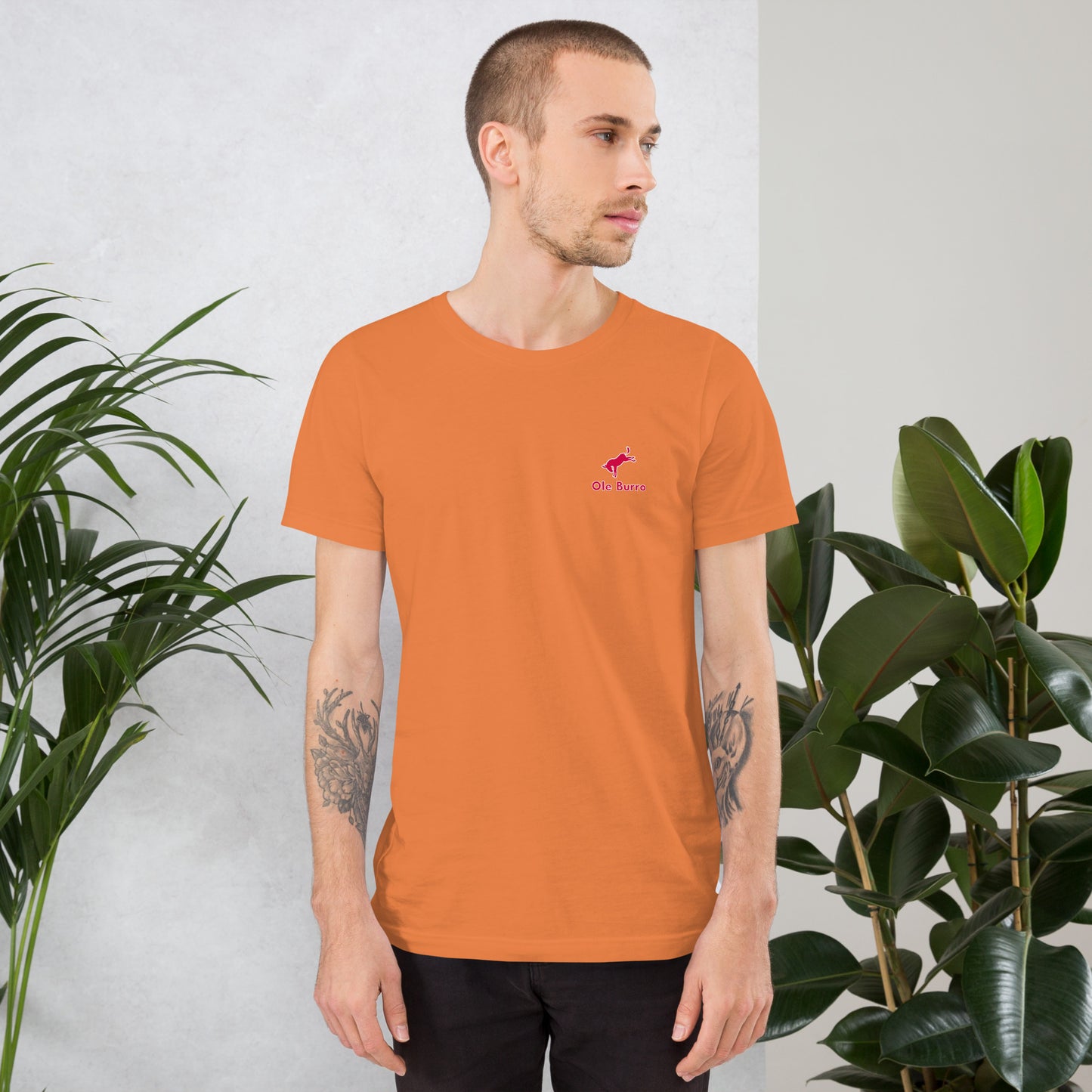 Man wearing an orange t-shirt with a small logo, standing against a light gray background with plants.