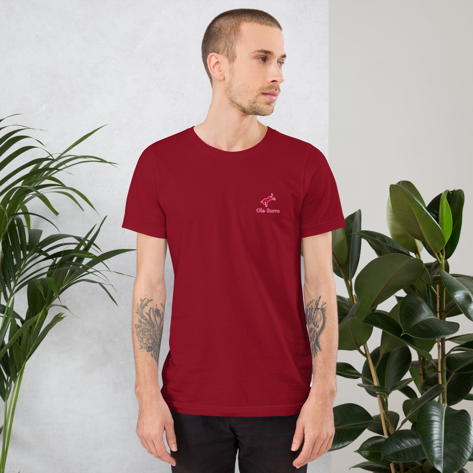 Man wearing a red t-shirt with a logo, standing between two plants against a light gray wall.
