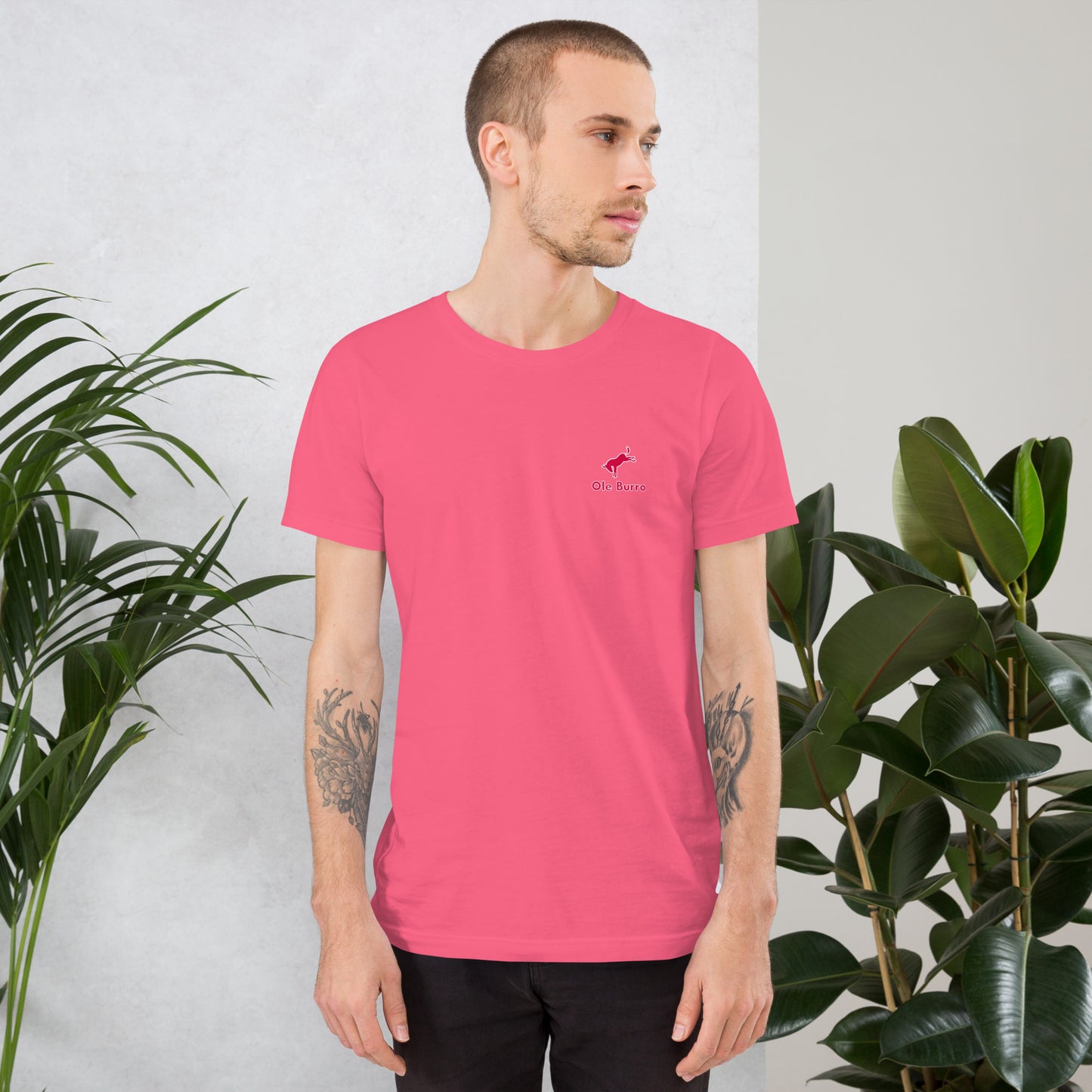 Man wearing a pink t-shirt with a small logo, standing against a light gray background with plants.