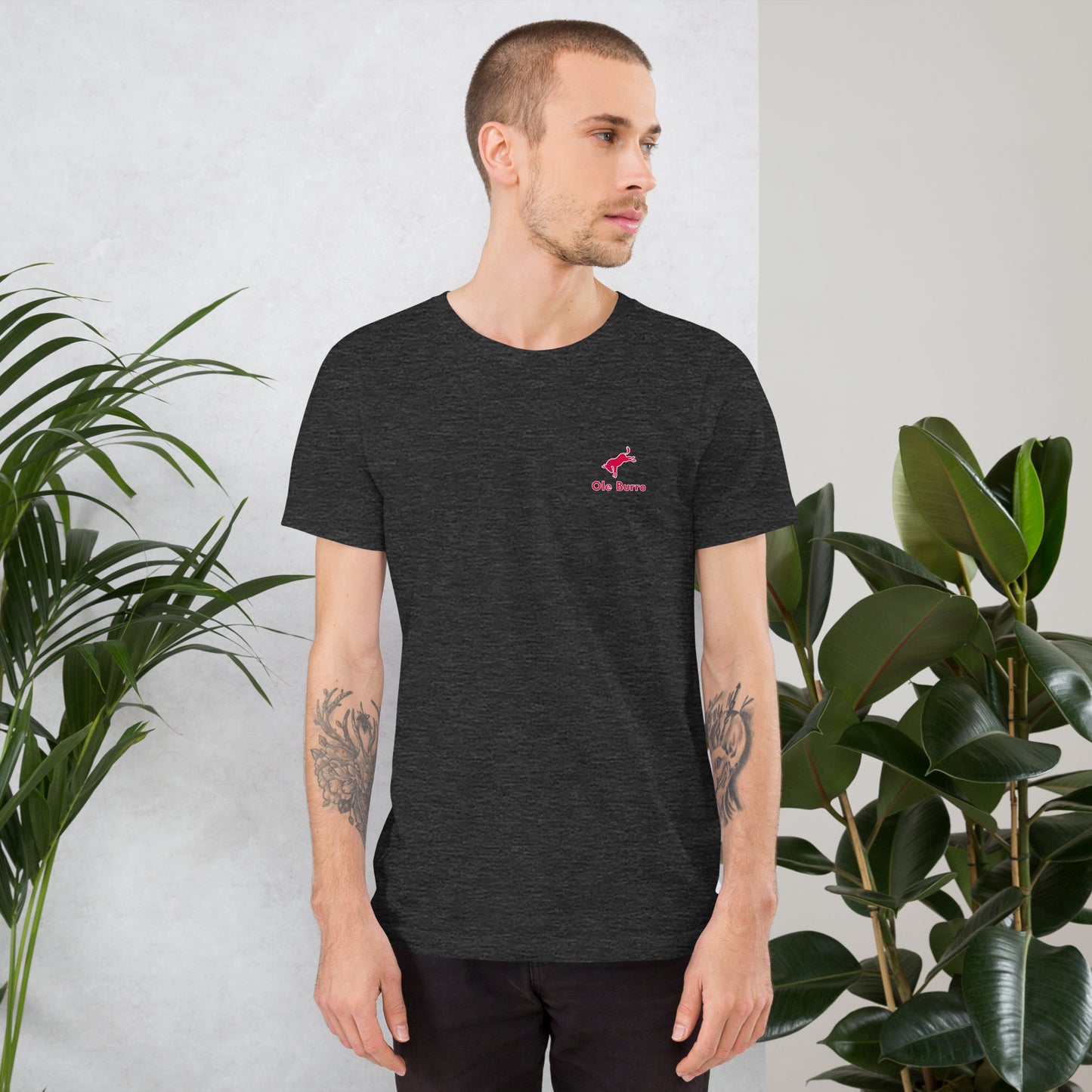 Man wearing a black t-shirt with a red logo, standing between two plants against a light gray wall.