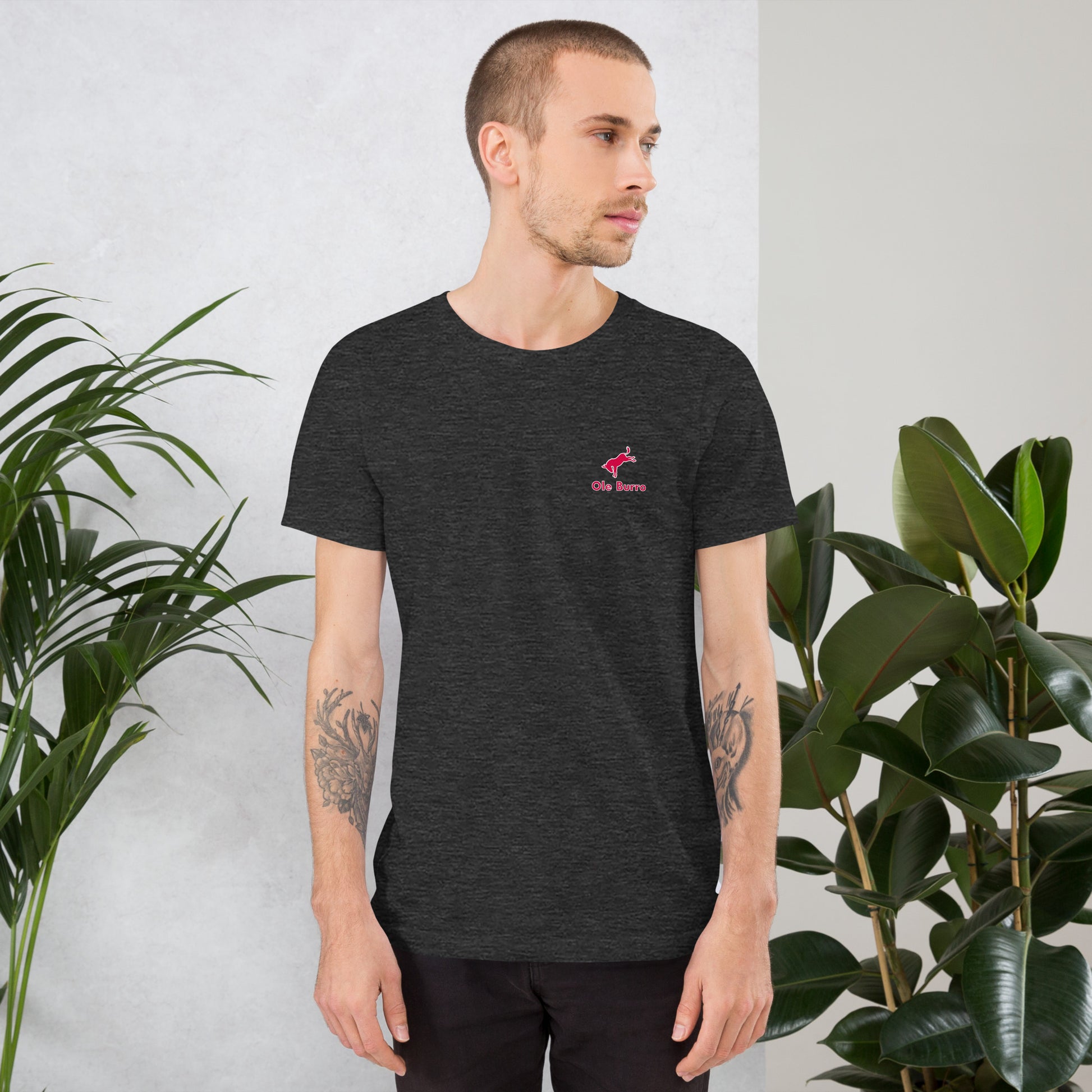 Man wearing a black t-shirt with a red logo, standing between two plants against a light gray wall.