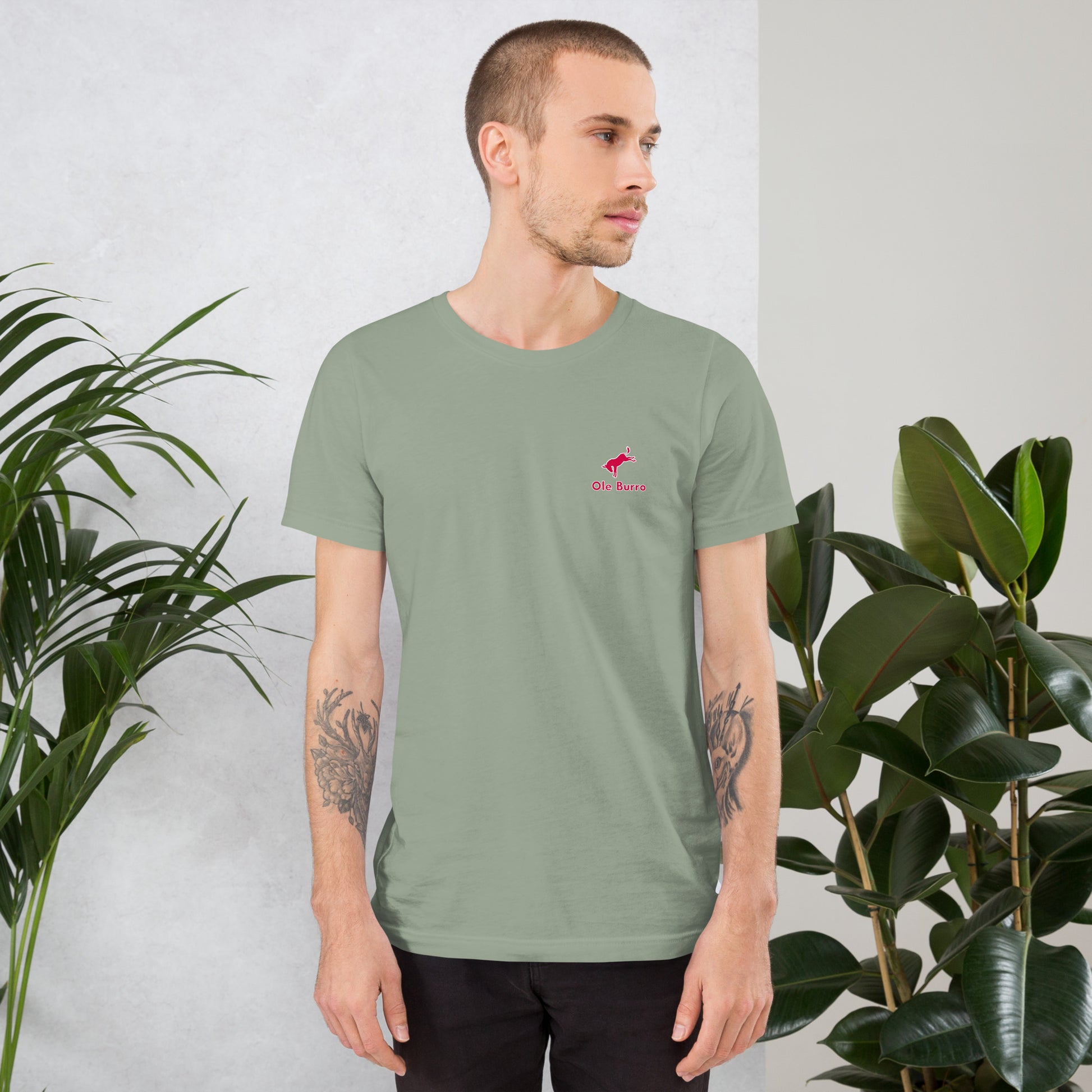 Man wearing a green t-shirt with a small red logo, standing between two potted plants.