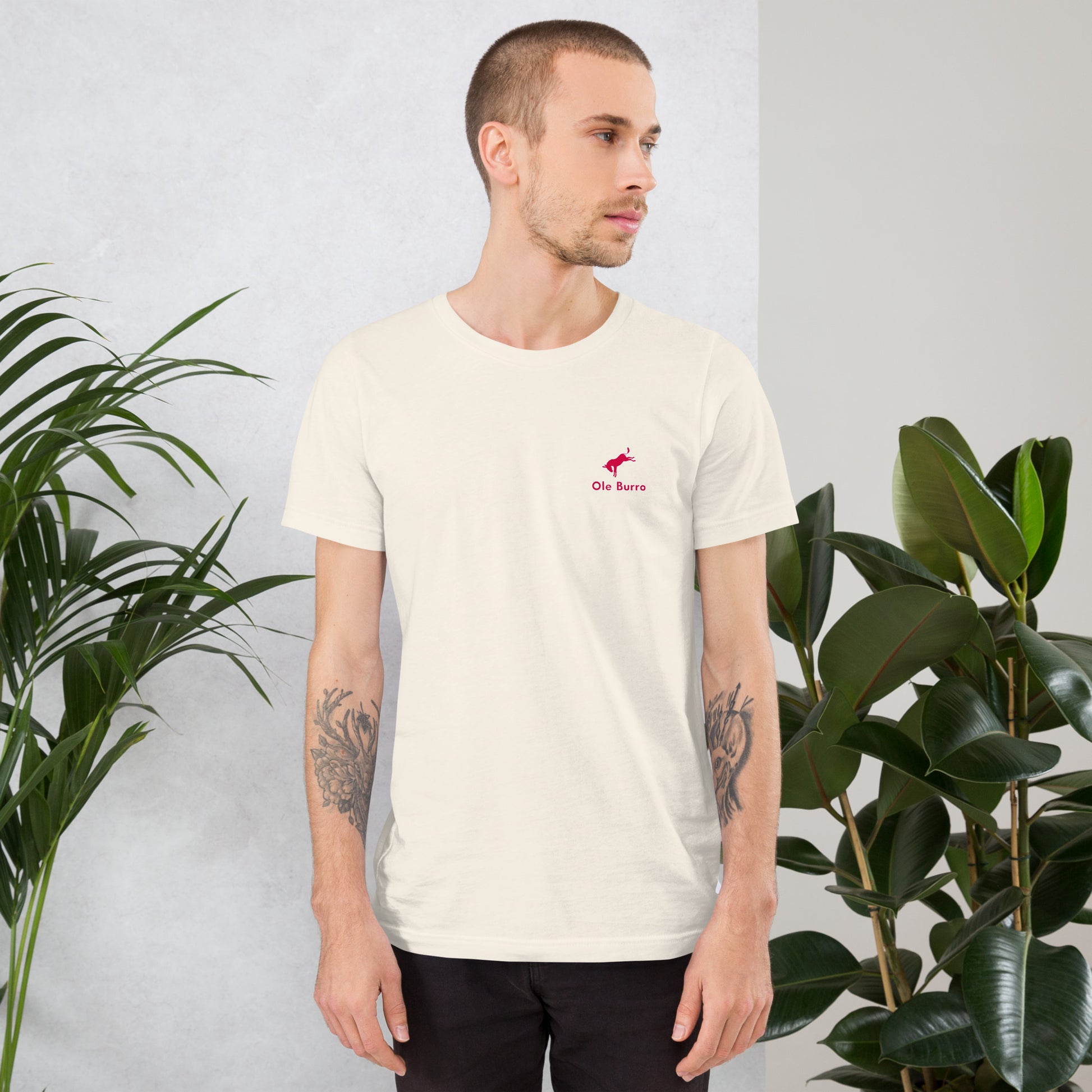 Man wearing a white t-shirt with a red logo, standing between two potted plants against a light gray wall.